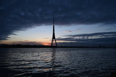 Büyük bir nehrin önü dalgalarla dolu, gün batımında dramatik mavi gökyüzü ve turuncu aksanlı. Nehrin karşı yakasında yer alan Riga TV kulesinin görüntüsü. Kuşlar uçuyor.