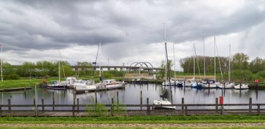 Sakin bir yat limanının üzerinde fırtınalı bir gökyüzü ve kıyıda yansımaları. Arka planda, Str Nehri üzerinde bir otoyol köprüsü var. Itzehoe. Almanya.