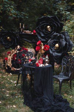 Moody outdoor Halloween scene with oversized black roses, a skull centerpiece, red roses and ornate vintage props on a draped table. Perfect for seasonal campaigns, event posters and spooky editorial use.
