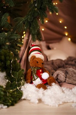 Çizgili şapka ve kırmızı eşarp takan kahverengi ayıcık çam dalları ve yapay karların arasında oturmuş, elinde kartopu tutuyordu. Arka planda sıcak peri ışıkları.