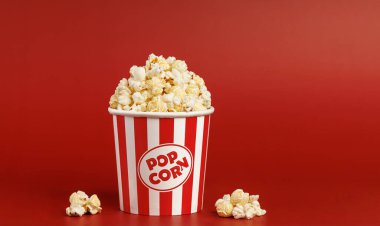 Çizgili bir kovanın yanına dökülen lezzetli patlamış mısır tanelerinin yüksek kaliteli görüntüsü. Eğlenceyi, sinema atmosferini ve boş zamanı temsil eden koyu kırmızı bir arkaplan..