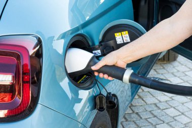 Beyaz bir kadının eli, mavi bir elektrikli aracın konektörüne bir CCS şarj prizi yerleştiriyor.