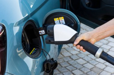 Beyaz bir kadının eli, mavi bir elektrikli aracın konektörüne bir CCS şarj prizi yerleştiriyor.