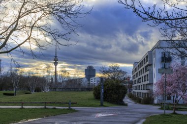Gün batımında güneş ve bulutların dramatik bir karışımı televizyon kulesi ve Milbertshofen Almanya Münih 'teki Petuelpark' taki BMW şirket binası manzarası 16.3.2024