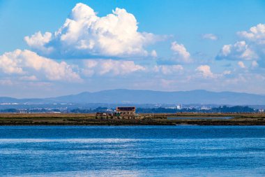 Sakin mavi suları ve dramatik bulutların altındaki uzak dağlarla kıyı şeridinde uzak bir bina..