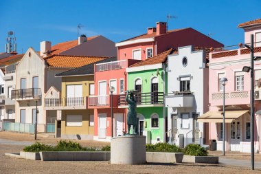 Sakin bir kasaba meydanında, Portekiz, Torreira, 13 Ekim 2025 'te berrak mavi gökyüzünün altında küçük heykeli olan renkli evler.