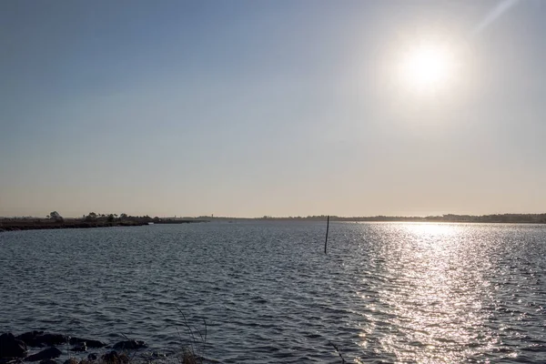 Parlak öğleden sonra güneşi uzak kıyı şeridi olan sakin göl sularını yansıtıyor, Portekiz, Q.ta das Ricos, 9 Ekim 2025