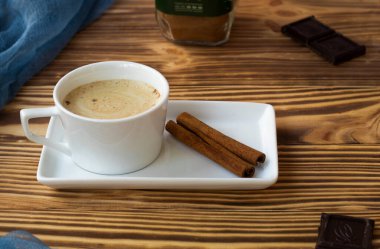 Bir fincan köpüklü latte, ahşap bir masada, kare bir tabağın üzerinde duruyor..