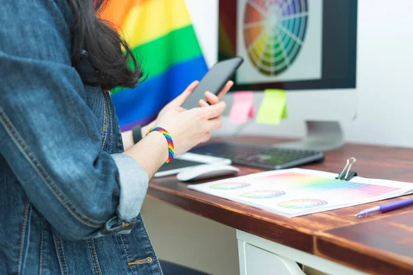 Woman in office with cell phone and LGBT accessories and gay flag. LGBTQIA culture.