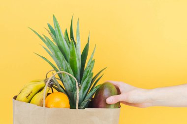 Sarı arka planda kadın eli olan tropik meyvelerle dolu kağıt alışveriş torbası. Boşluğu kopyala.