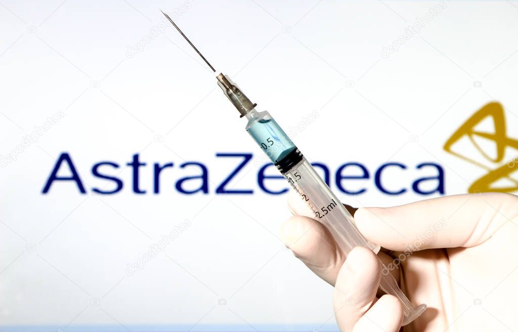 Madrid, Spain- January 7, 2020: Hand on white surgery glove holding Syringe with Covid vaccine. In the background Astrazeneca Laboratory logo on white background.