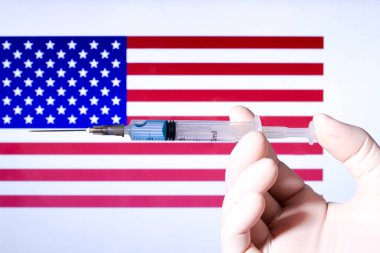 Ellerinde şırınga ve kovid aşısı olan eldiven var. Arka planda ABD bayrağı