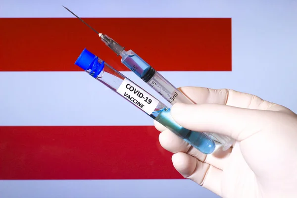 Hand in surgery glove holding syringe with covid vaccine. Austrian flag in the background