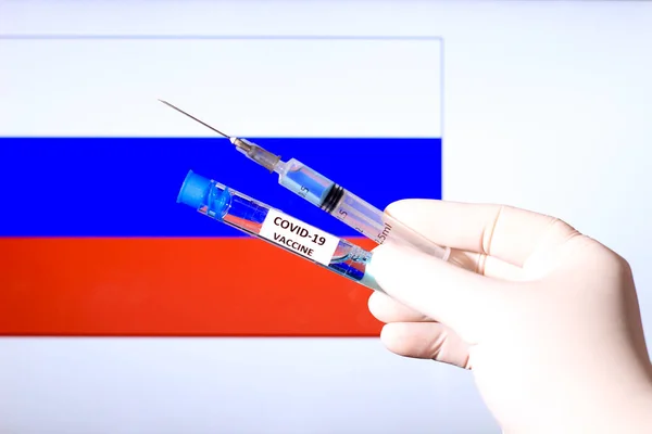 Hand in surgery glove holding syringe with covid vaccine. Russian flag in the background