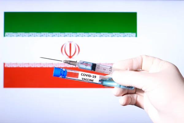 Hand in surgery glove holding syringe with covid vaccine. Iranian flag in the background