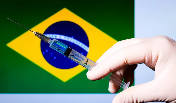 Hand in surgery glove holding syringe with covid vaccine. Brazilian flag in the background