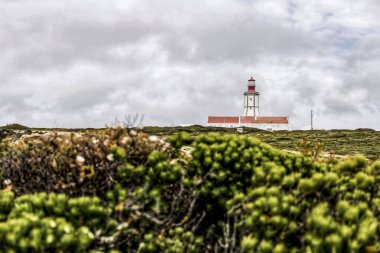 Uçurumun tepesinde deniz feneri ve Portekiz Cape Espichel 'de bulutlu gökyüzünün altında bitki örtüsüyle çevrili.