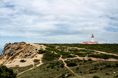 Uçurumun tepesinde deniz feneri ve Portekiz Cape Espichel 'de bulutlu gökyüzünün altında bitki örtüsüyle çevrili.
