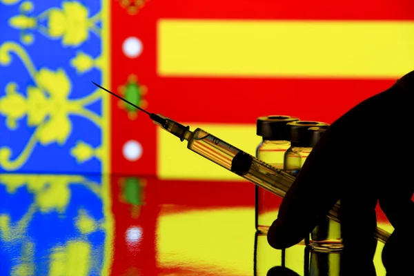 Hand in surgery glove holding syringe with covid vaccine. Valencian Community flag in the background