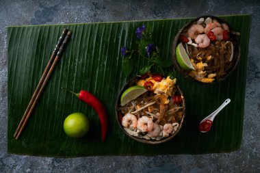 Geleneksel Tayland Eriştesi. Kızarmış Noodles Pad Thai, karides ve hindistan cevizi kabında tavuk taze limonlu, muz yaprağının üzerinde kırmızı biber, ahşap sandalyede. Yan görüş. Seçici odak 