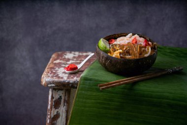 Geleneksel Tayland Eriştesi. Kızarmış Noodles Pad Thai, karides ve hindistan cevizi kabında tavuk taze limonlu, muz yaprağının üzerinde kırmızı biber, ahşap sandalyede. Yan görüş. Seçici odak 