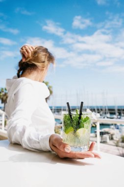 Açık hava barında bir bardak mojito kokteyliyle marina manzarasının tadını çıkaran bir kadın. Seçici odak. 