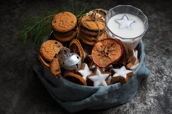 A selection of different cookies (star glazed cookies, waffles and chocolate chip cookies) in a box served with galass of milk decorated with Christmas toy and fur tree branch.