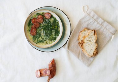 Yeşil sebzeli Caldo Verde Çorbası ve seramik tabağın üzerine seramik kasede dilimlenmiş sucuk. Pamuk ve sucuk yerine ekmek parçaları. Üst Görünüm
