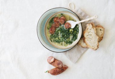 Yeşil sebzeli Caldo Verde Çorbası ve seramik kasenin üstünde seramik tabak üzerinde seramik kaşık ile servis edilmiş ekmek ve sucuk parçaları. Üst Görünüm