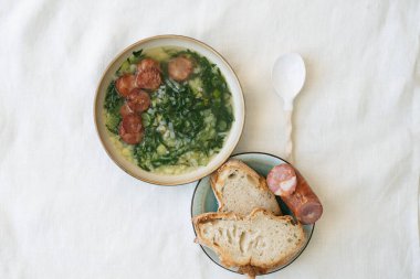 Yeşil sebzeli Caldo Verde Çorbası ve seramik kaşıkla seramik kasenin tepesinde doğranmış sucuk. Seramik tabakta ekmek ve sucuk parçaları. Üst Görünüm