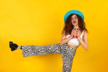 Beyaz güneş gözlüğü takan ve kırmızı kokteyl içerken eğlenen yaz kıyafetli bir kız. Sarı arkaplanda poz.