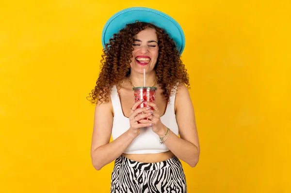 Girl in summer outfit wearing white trendy sunglasses and enjoying while drinking a red cocktail. Posing on the yellow background.