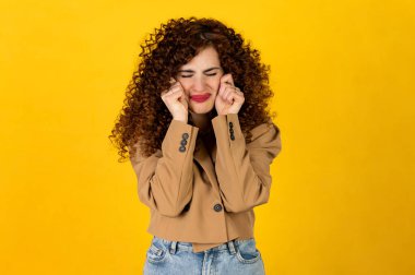 Stüdyoda sarı arka planda poz veren kot ve ceketli kıvırcık kız. Üzgün kadın yüz ifadesi. Ağlıyor ve ellerinle yüzünü ovuşturuyorsun..