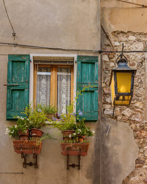 Typical old Italian facade window with green shutters and flowers in planters ,an old lantern on the wall