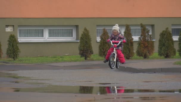 Kid försöker sitta på en cykel Wheeling liten flicka Rider två hjul cykel av en skola gården gräsmatta vintergröna träd våta vägen pölar vinter eller höst — Stockvideo