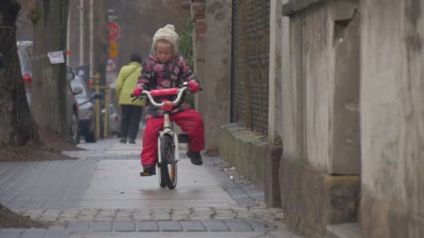 어린 소녀는 카메라 를 향해 자전거를 타고 분홍색 자전거에 앉아 작은 소녀는 도시 거리 걷는 사람들 나무에 의해 자전거 이륜 자전거를 타고 — 비디오