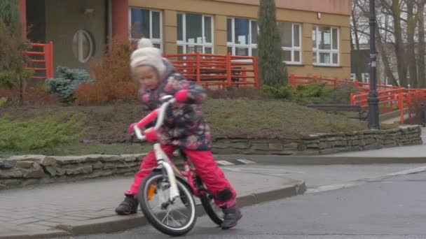 Kid börjar hjulet cykeln gör ett försök att försöka hård liten flicka rider en cykel tvåhjuliga cykel av en stadsgata byggnad vintergröna träd — Stockvideo