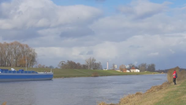 Mann läuft Touristin dreht um und läuft auf Kamera blaues Schiff auf einem kleinen Fluss Fabrik Rauch aus einem Rohr am gegenüberliegenden Ufer graue Kumulus treiben — Stockvideo