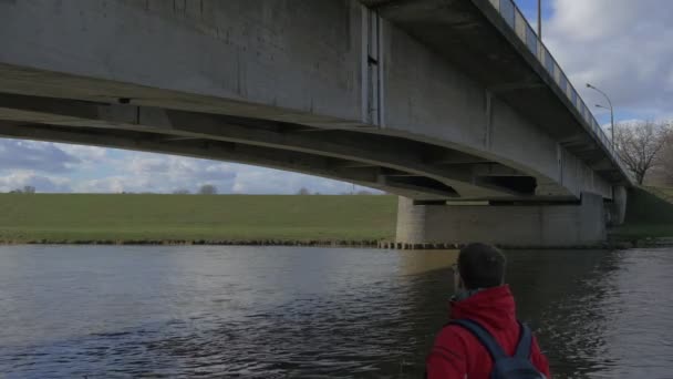 Mann blickt auf betonierte alte Brücke Wasser fließt unter Brücke grüne Bank kahle Äste Bäume am gegenüberliegenden Ufer Himmel Spiegelung in einem plätschernden Wasser bewölkt — Stockvideo