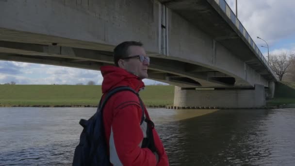 El hombre está parado en el viejo puente de hormigón que mira alrededor de los flujos de agua debajo del puente Banco Verde Ramas desnudas Árboles en el banco opuesto Reflexión del cielo en una ondulación — Vídeos de Stock