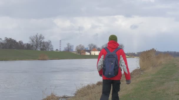 Un touriste marche le long d'une rivière Backpacker en veste de sport rouge s'arrête et regarde l'eau tournant la tête Clousy Day Opposite Bank — Video