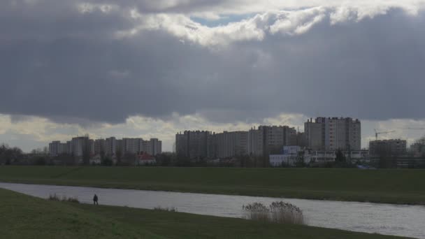 Die Silhouette des Mannes läuft am grünen Flussufer entlang, wo mehrstöckige Wohnhäuser am gegenüberliegenden Ufer stehen, am Horizont schweben himmelgraue Wolken. — Stockvideo