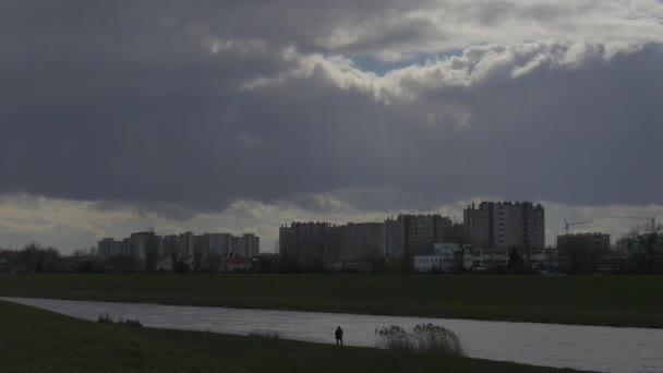 Man 's Silhouette is Walking by River Bank Green Grass Residental Multi-Storeyed Houses on Opposite Bank on a Horizon Lagre Piece of Sky Gray Fouds — стоковое видео