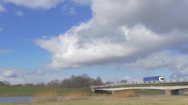 Blauwe vrachtwagen auto's en vrachtwagens in zonnige dag gedreven door een brug van de auto door de kleine rivier witte Cumulus zweven door blauwe lucht, groene weides verlicht met zon — Stockvideo