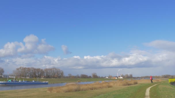 Mann in Rot fernab von Kamera-Backpacker läuft entlang eines Flusses an einer Wiese stehend für eine Weile grüne Grasgebäude am Horizont weiße Wolken blauer Himmel — Stockvideo