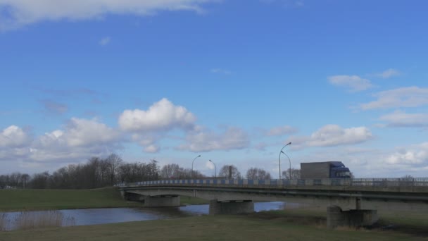 Λεωφορείων επιβατηγά αυτοκίνητα και τα φορτηγά οδηγούνται γέφυρα μέσω μικρού ποταμού επιπλέουν πυκνό λευκό σύννεφο από μπλε ουρανό πράσινα λιβάδια γυμνοί κλάδοι δέντρα — Αρχείο Βίντεο