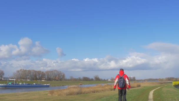 Turist är gångavstånd härifrån längs en flod av ängen Backpacker i röd jacka på en Green Grass byggnader på en horisont vita moln på en blå klar himmel — Stockvideo