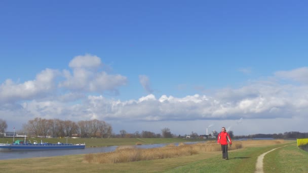 バックパッカーは、歩くに沿って、川に向かってカメラ草原緑草建築物赤メガネでブルーの明確な空男の地平線白い雲に — ストック動画
