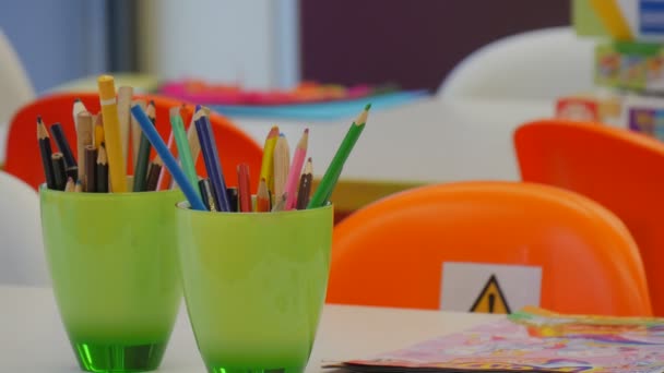 Twee groene Cups met kleurrijke potloden op een tafel iemand zet een dozen papier op een tafel oranje stoelen tafels Violet muren Windows van een klaslokaal — Stockvideo