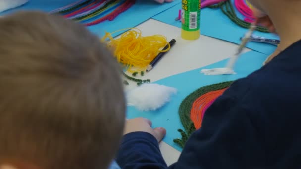 Kid je lepení mraky bavlny na modrý papír mraky na obloze dětské ruce dělají Rainbow z barevné vlákna sedící u stolu učebny — Stock video